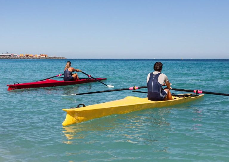 X-Adventure coastal rowing (gelbes & rotes Ruderboot) Zwei Ruderer in Booten auf ruhigem Wasser unter klarem Himmel.